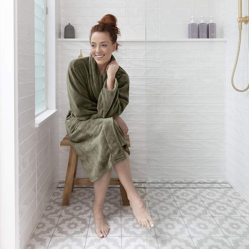 alt="A woman wearing an olive green microplush robe sitting on a chair in the bathroom about to take a bath"
