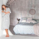 alt="A woman wearing a silver microplush robe in the morning beside her bed"