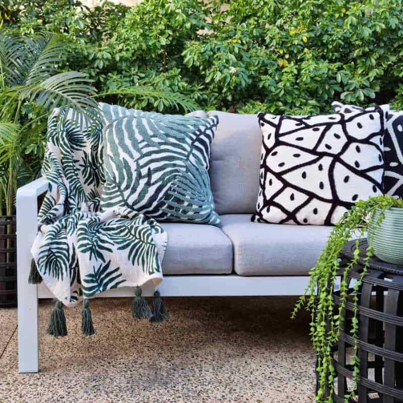 alt="Coastal-inspired cushion with double-sided foliage design, placed on a sofa in an outdoor space"