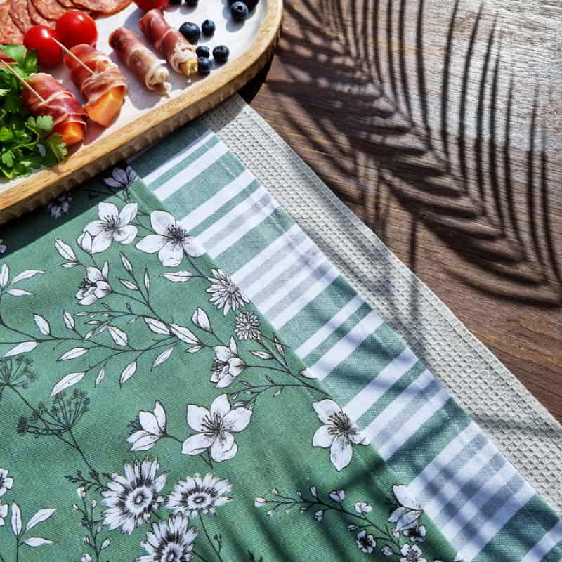 alt="Set of green tea towels feature one printed design, one striped design and one plain waffle in a table.