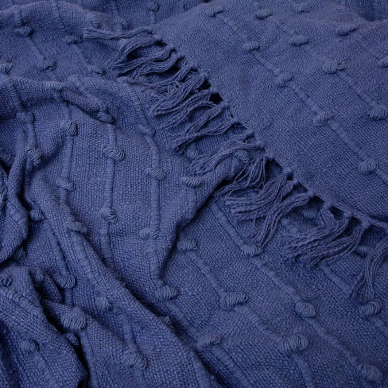 alt="Close up look of a textured solid colour throw with striped and looped stitching, adorned with tassels, in Blueberry hue."