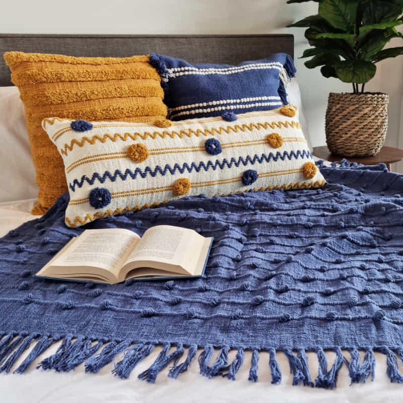  alt="Textured solid colour throw with striped and looped stitching, adorned with tassels, in Blueberry hue in a cosy bedroom."