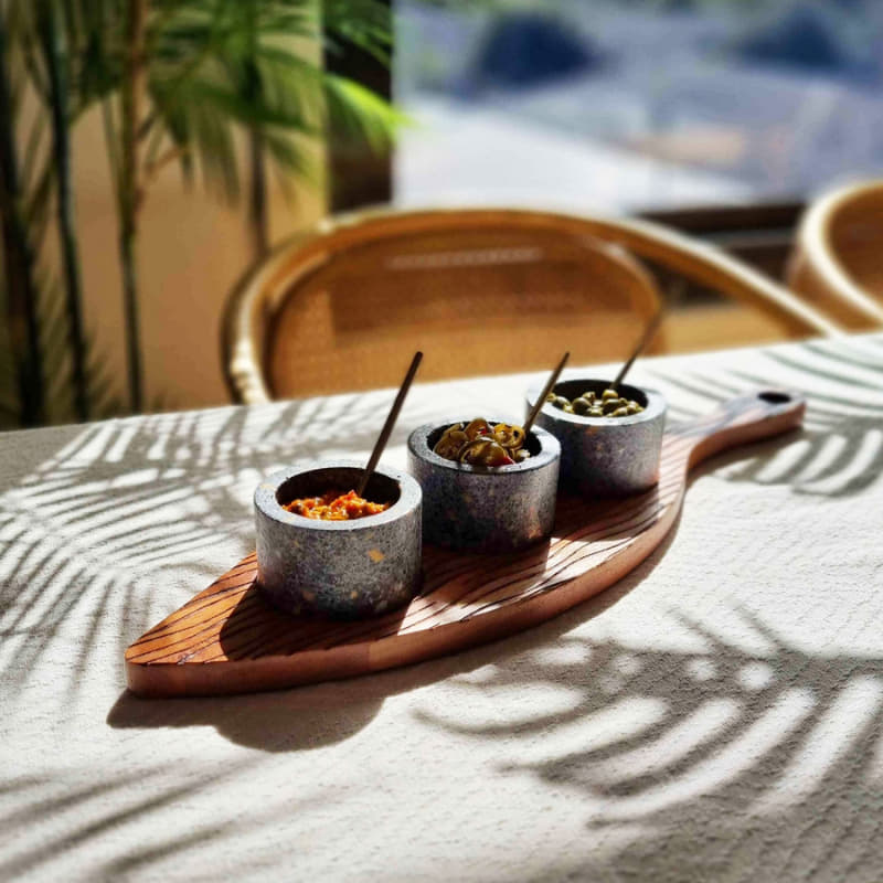alt="A serving board with condiment bowls and spoons features three handcrafted detachable terrazzo bowls nestled into a beautifully carved acacia wood tray, each with its own golden spoon in a table."